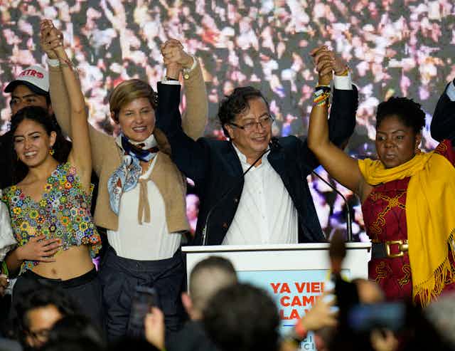 A man stands holding raised hands with two women on each side of him before a crowd of people.