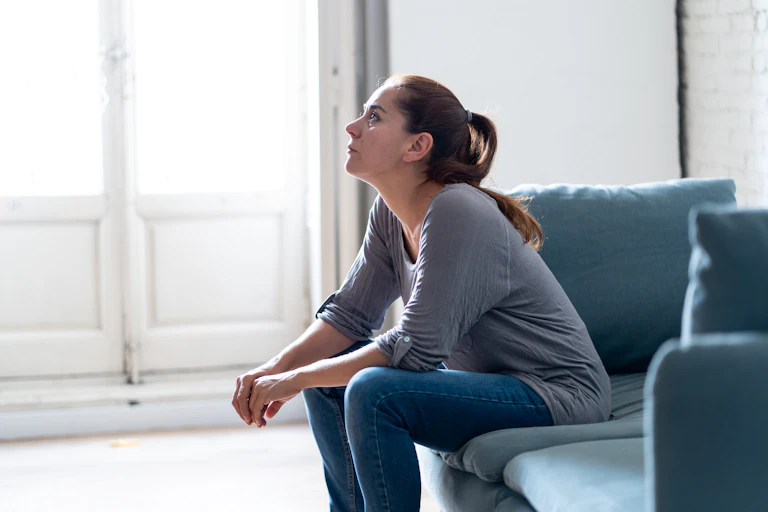 Young woman with long brown hair in a ponytail sits on a sofa and stares into the distance