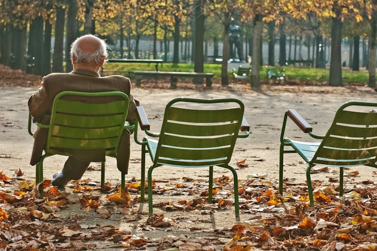 Older man sits alone in a green chair at a park, autumn leaves on the ground