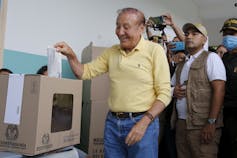 A man in a yellow polo shirt and jeans pushes his ballot into a ballot box.