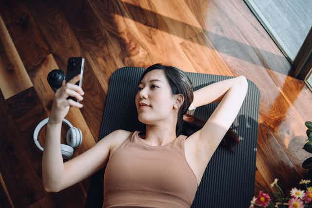 High-angle shot of a young Asian woman relaxing on a yoga mat and looking at her smartphone, with a reusable water bottle and headphones by her side.