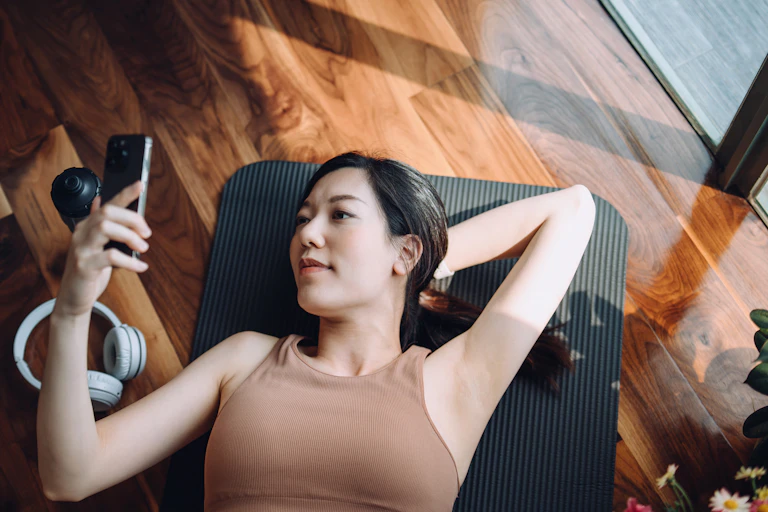 High-angle shot of a young Asian woman relaxing on a yoga mat and looking at her smartphone, with a reusable water bottle and headphones by her side.