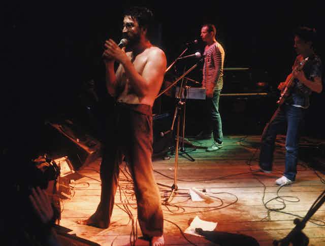 Shirtless man sings into a mic on a stage with bandmates in the background.
