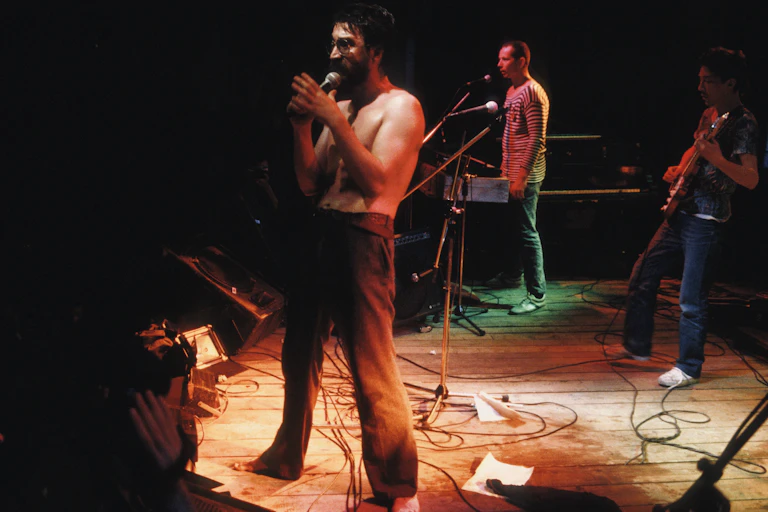 Shirtless man sings into a mic on a stage with bandmates in the background.