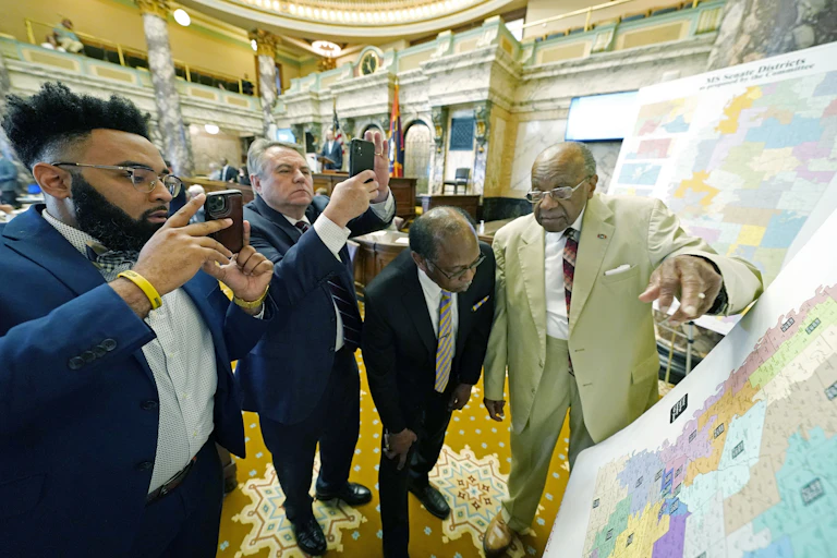 Four men in suits in a fancy large public chamber looking at large maps of the state.
