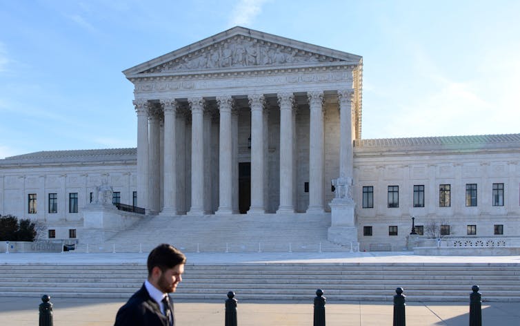 A large, elegant white building with columns and a plaza in front of it with one man walking near it.