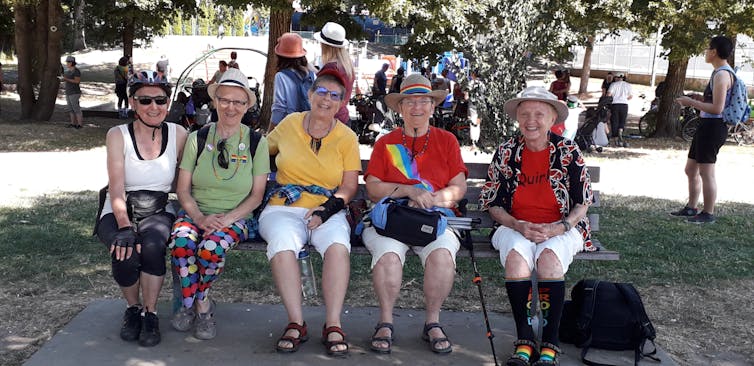 A group of older women sit on a bench donned in Pride attire.