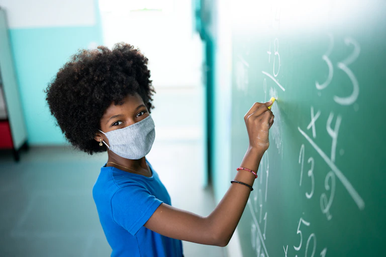 A school girl writing on the blackboard in a classroom. She's wearing a face mask.