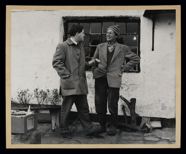 Black and white photo, one man mock interviews an older man with a tape recorder and old-fashioned microphone, standing outside a rural home. The older man is wearing a knit cap and smiling