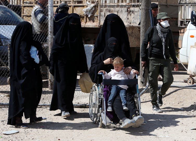 Three women in black burqas assist another woman in a wheelchair, who is holding a child on her lap.