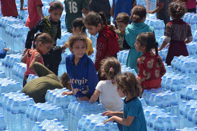 Children stand between rows of packaged bottles of water.