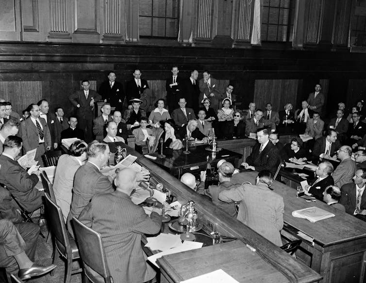 A crowded room where a handful of men are sitting at a raised desk, while one woman talks in the audience