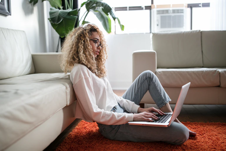 Woman sits on her living room floor, researching on her laptop.