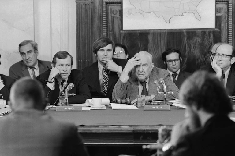 A handful of men in suits at a desk, listening to someone in front of them giving testimony.