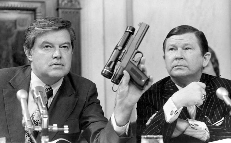 Two men sitting at a table, one holding up an oddly shaped gun.