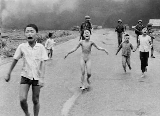 Frightened and tearful children, including one without clothes, run down a road in front of soldiers.