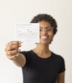 Woman smiling outside at attention raises her hand holding vaccination record card