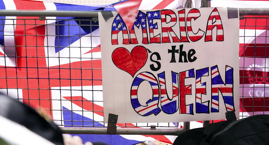 A placard reading "America hearts the Queen" is attached to a security barrier in front of a British and American flag.