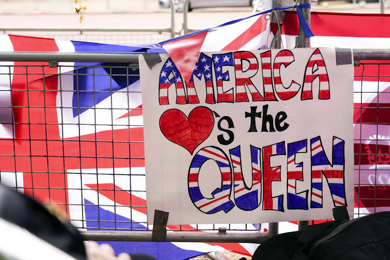 A placard reading "America hearts the Queen" is attached to a security barrier in front of a British and American flag.