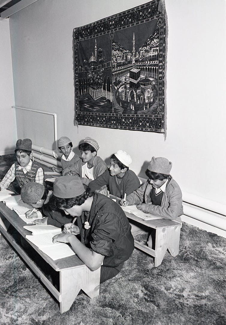 Young boys wearing prayer caps read and write at low wooden desks while seated on a carpeted floor.