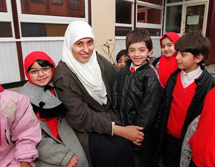 Smiling school pupils in grey and red surround a woman in a white headscarf.
