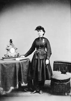 An old black and white photograph depicts a white woman wearing a dark hat and dress in a formal pose next to a table.