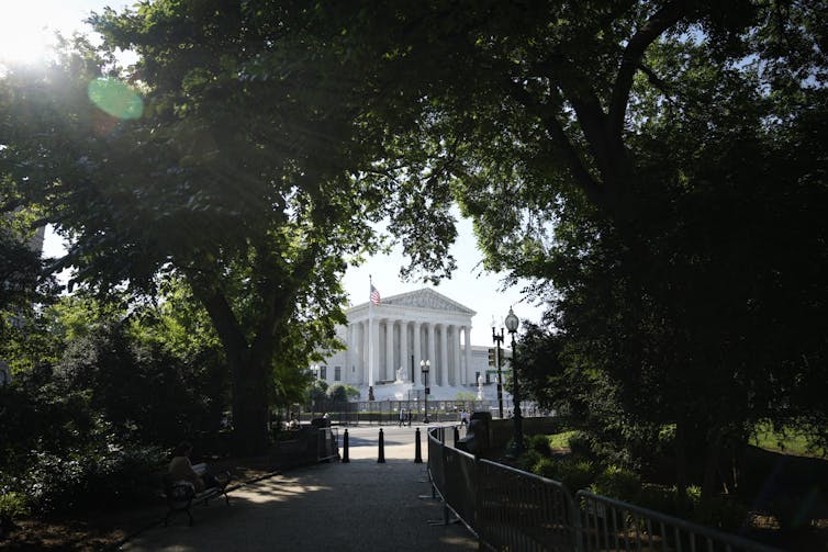The Supreme Court is seen through an opening among the trees on a shady road.