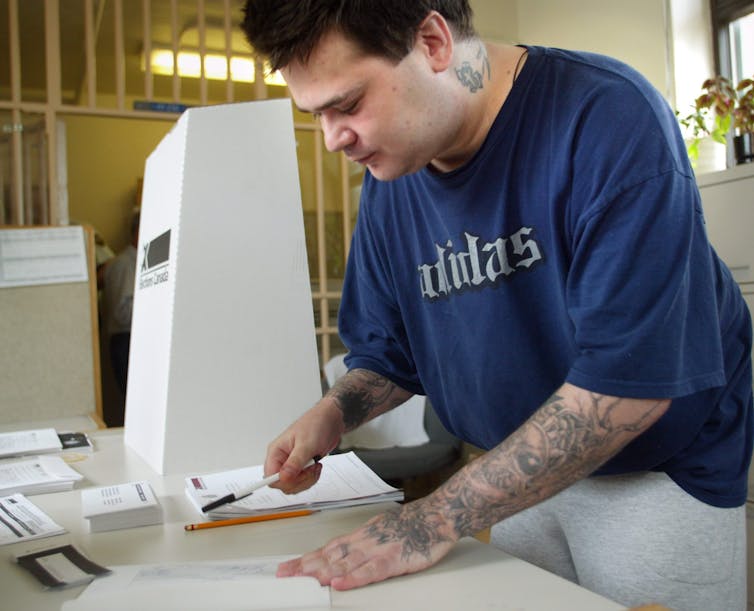 A man in a T-shirt and sweatpants leans over a table to fill out paperwork with a pen
