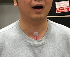 Cropped image of a man in a gray T-shirt with a small round sensor patch on his neck just above the neckline of his shirt