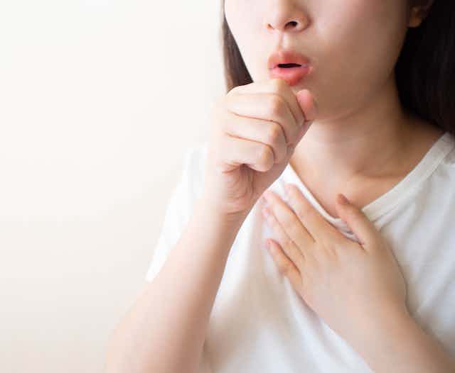 Cropped image of a woman in a white T-shirt with her hand on her chest and her hand in front of her mouth as though coughing
