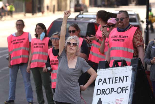 A woman in gray raises up her hand, in front of a row of people wearing pink vests that say 'clinic escort volunteer.'" 
