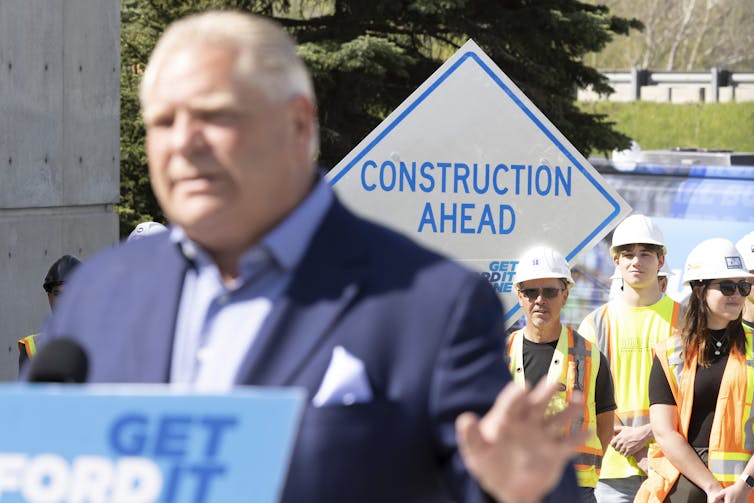 Doug Ford stands at a podium with a sign behind him that says Construction Ahead and construction workers.