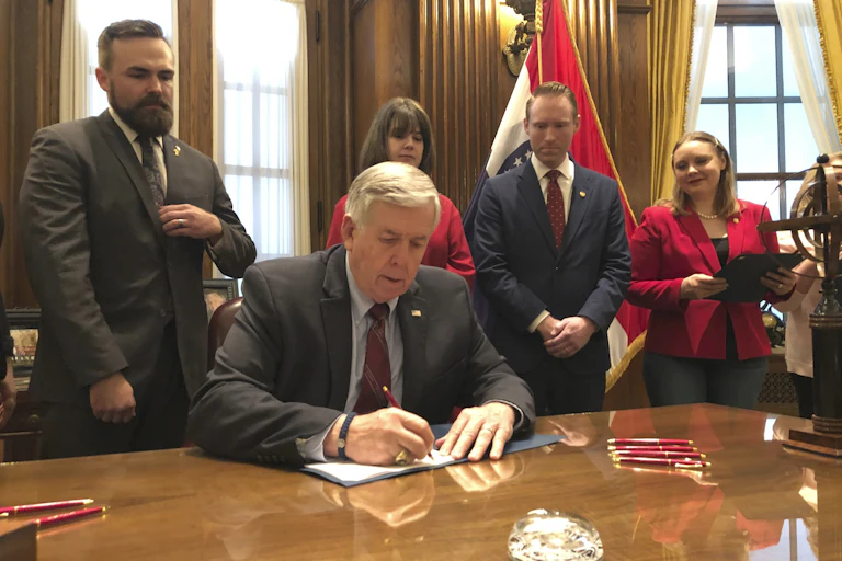 A man at a desk signs a document with several people watching