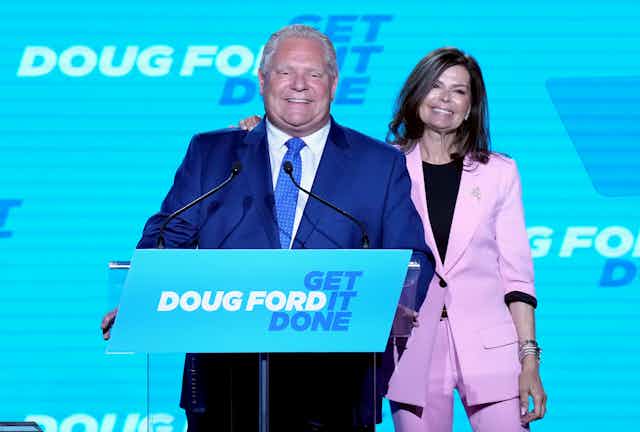 A smiling Doug Ford stands in front of a podium that says Doug Ford Get It Done while his wife Karla puts her arm around him.