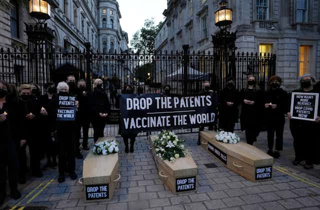 Protesters dressed in black with three replica coffins on the ground labelled 'Drop the Patents' and a banner reading 'Drop the patents vaccinate the world' in front of an ironwork gate