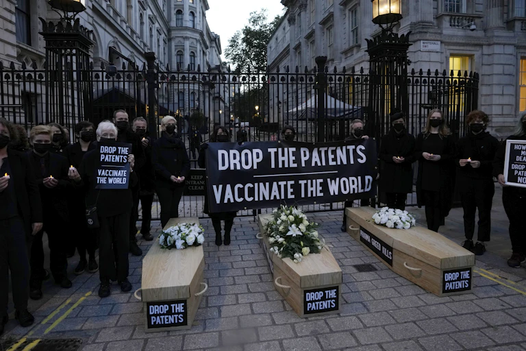 Protesters dressed in black with three replica coffins on the ground labelled 'Drop the Patents' and a banner reading 'Drop the patents vaccinate the world' in front of an ironwork gate
