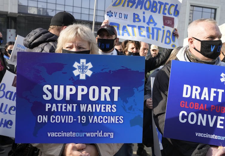 A crowd outdoors with people holding blue signs reading'Support patent waivers on COVID-19 vaccines'