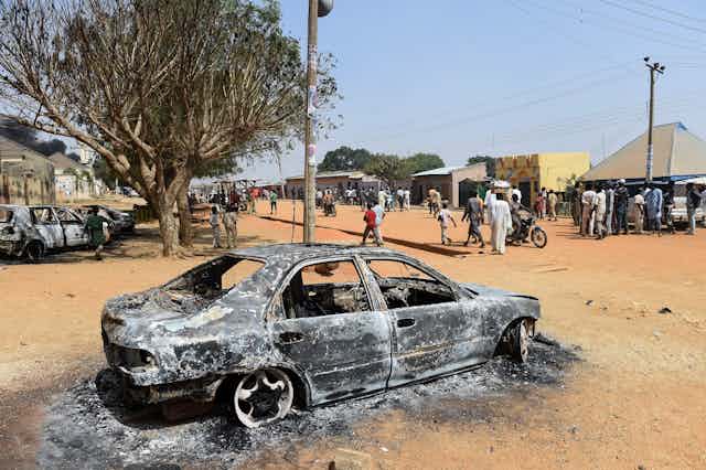 Burnt out car on an unpaved road, with buildings and people in the background