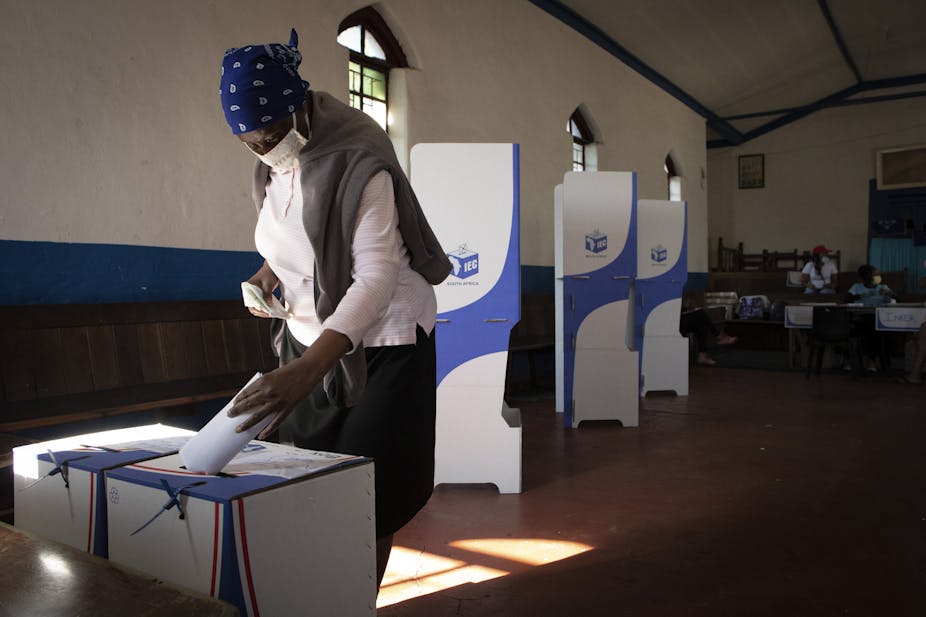 A woman wearing a headscarf and a COVID mast puts a ballot paper in a ballot box.