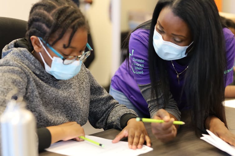 A teacher at a desk helps a teenaged female student with her work.