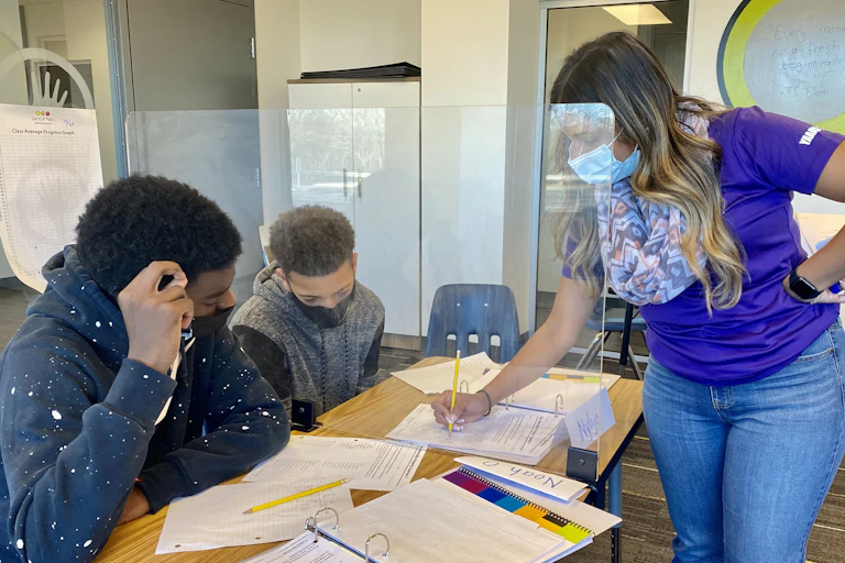 Two students are seen at a desk in face masks while a teacher holding a pencil writes on a sheet of paper.