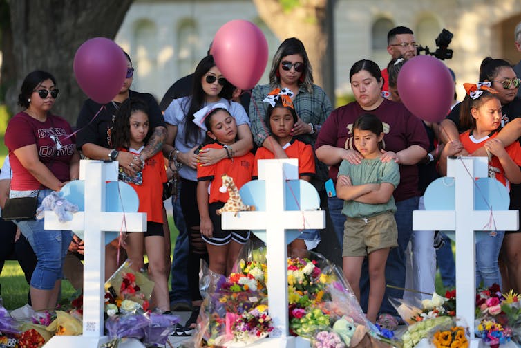 Adults stand behind children, holding balloons, in front of white cross memorials.
