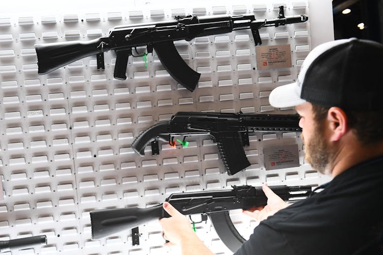 A man holds an AK-47, picking one off a white wall with other guns