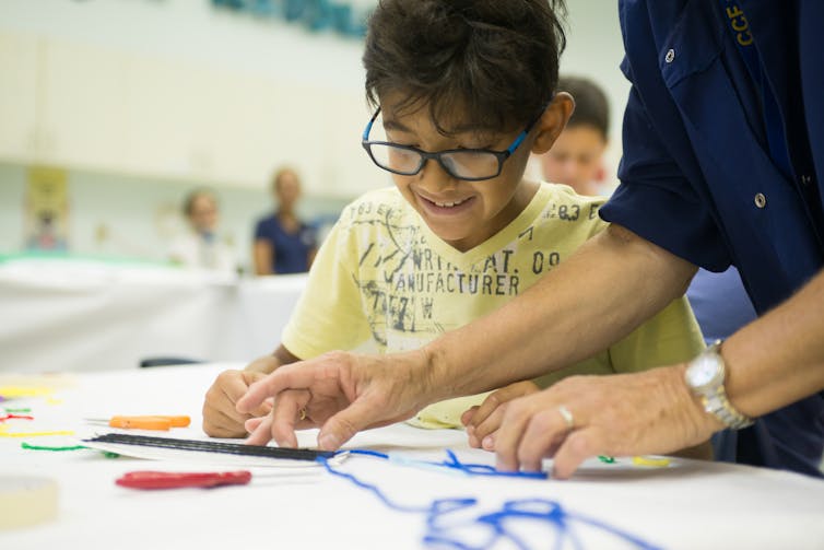 A child is seated a table working on a project as a teacher stands nearby.