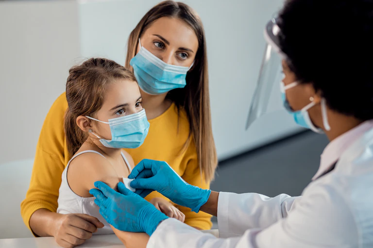 A health-care worker applies a bandaid to the upper arm of a young girl, who is sitting with her mother.