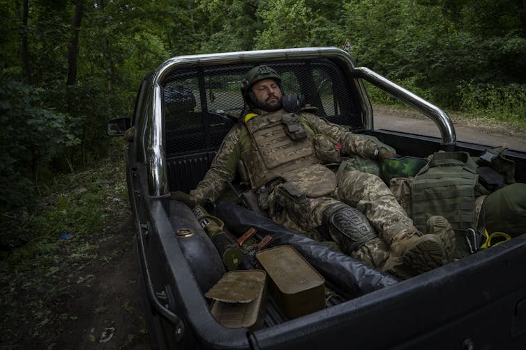 A soldier in full combat gear sits in the back of a military vehicle.