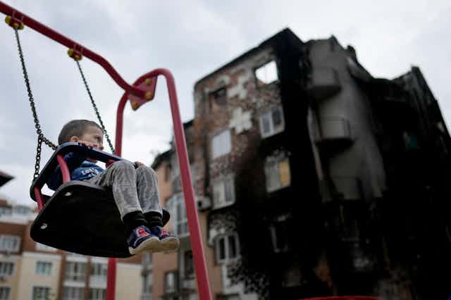 A boy sits in a swing outside a destroyed building.