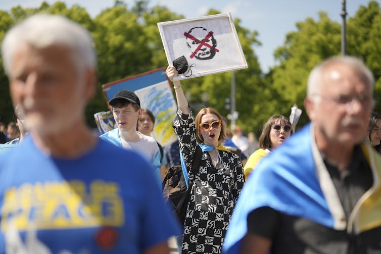 A woman in a checked coat holds up an anti-Putin sign behind men dressed in Ukrainian colours.