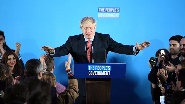 A smiling Boris Johnson addresses a clapping crowd from a lectern that says 'The People's Government'.