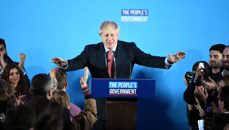 A smiling Boris Johnson addresses a clapping crowd from a lectern that says 'The People's Government'.